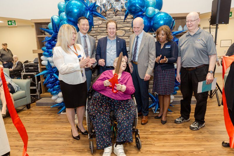 A woman in a wheelchair cuts a red ribbon at an indoor event, surrounded by six smiling adults and blue balloon decorations.
