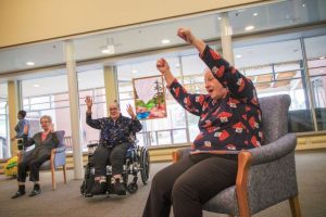 Three older adults, one in a wheelchair, sit indoors with arms raised in a group activity. Large windows and a colorful painting are in the background.
