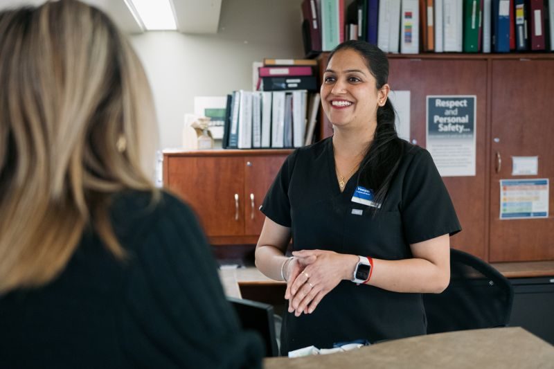 A woman in medical scrubs stands at a reception desk, smiling and talking to another woman whose back is to the camera. Shelves with files and safety signs are visible in the background.