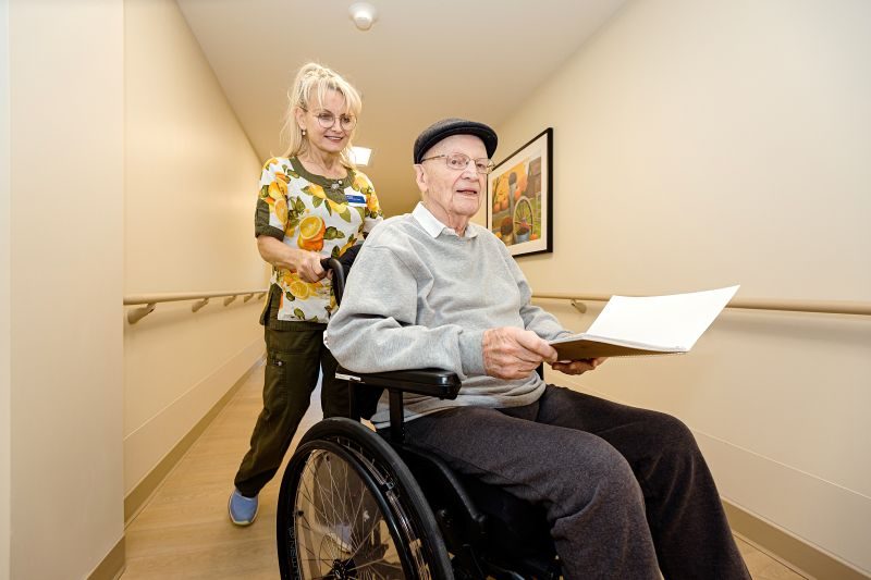 A woman pushes an elderly man in a wheelchair down a hallway; the man holds a folder and both appear to be in an assisted living facility.