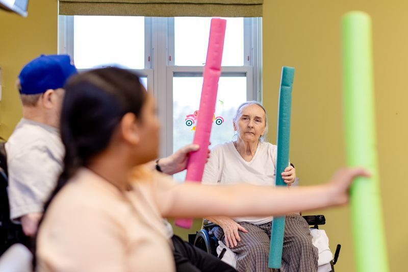 Elderly people sitting indoors participate in an exercise activity, each holding a colorful foam stick.