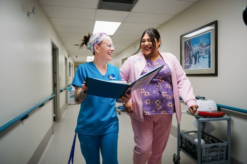 Two healthcare workers in scrubs walk down a hospital hallway, smiling and looking at a large binder together.