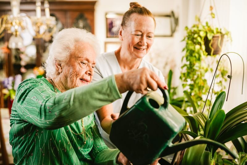 An elderly woman waters houseplants with a green watering can while another woman stands beside her, both smiling in a bright, indoor setting.