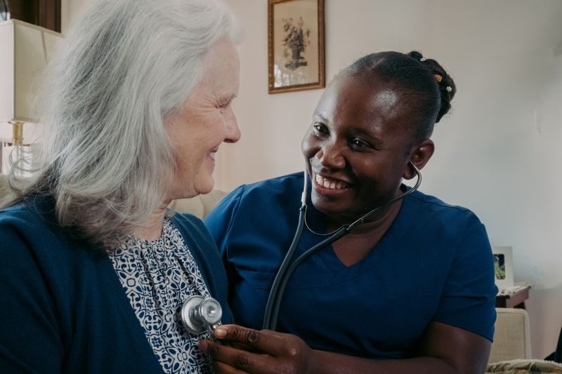 A healthcare worker in blue scrubs uses a stethoscope to check an older woman's heartbeat. Both are smiling in a home setting.
