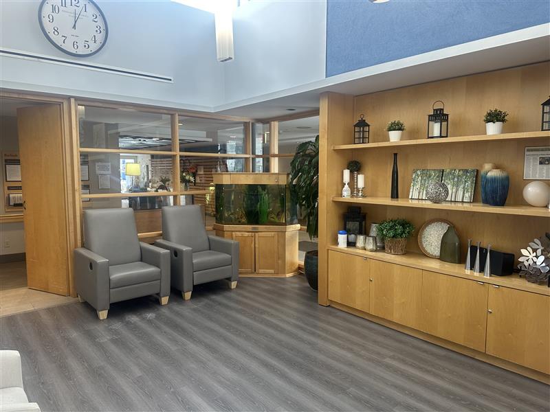 Modern waiting area with two gray chairs, a large fish tank, a wooden shelving unit with decor, and a wall clock above the seating.