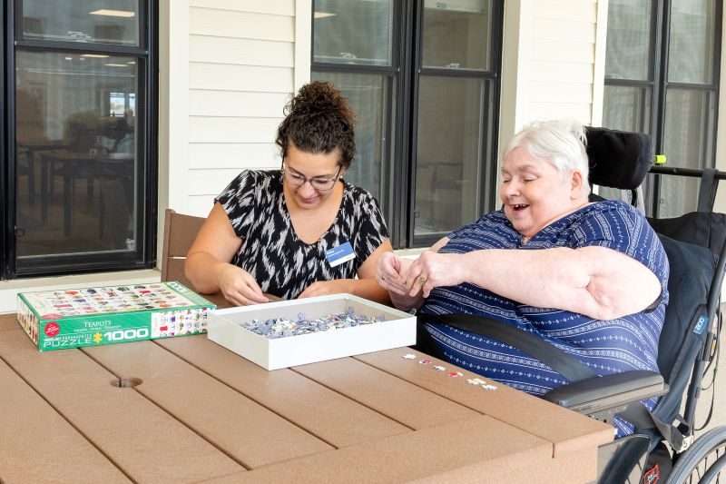 A woman in a wheelchair and another woman sit at a table assembling a jigsaw puzzle together indoors.