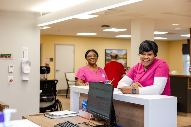 Two healthcare workers in pink scrubs stand behind a reception desk, smiling at the camera in a brightly lit medical facility.