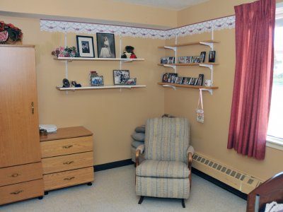 A small, tidy room with a patterned armchair, wooden dresser, shelves with photos and decorations, and a window with red curtains.