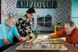 Two older men play a board game at a table while a younger woman stands between them, watching and smiling in a brightly decorated room.