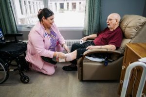 A healthcare worker kneels and bandages the leg of an elderly man sitting in a recliner in a care facility.