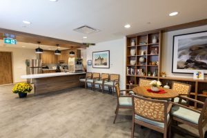 A modern lounge area with a round table and chairs, a built-in bookshelf, framed art, yellow flowers, and a kitchenette with coffee machines in the background.