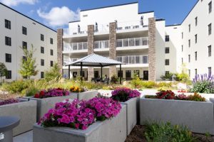 A modern apartment building courtyard with flower beds, a gazebo with seating, and landscaped greenery under a blue sky.