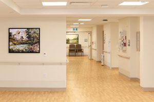 A clean, empty hospital hallway with beige walls and light wood floors, featuring a landscape painting and chairs at the end of the corridor.