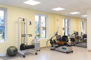 A bright gym room with exercise equipment, including a recumbent stepper, a weight machine, and an exercise ball, next to large windows letting in natural light.