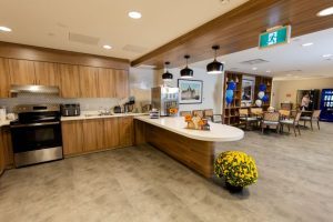 Modern communal kitchen and dining area with stainless steel appliances, wooden cabinets, a white island, potted yellow flowers, and blue balloons in the background.