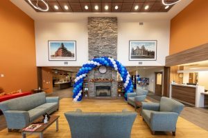A modern lobby with gray seating, a stone fireplace, a clock, blue and white balloon arch, two framed photos, and wood accents.