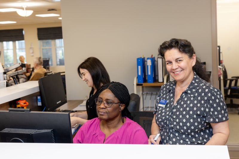 Three women are working at a reception desk with computers; one is seated in a pink shirt, another is standing and smiling, and a third is in the background typing.