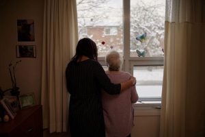 Two women, one younger and one older, stand by a window with butterfly stickers, looking outside at a snowy scene. The younger woman has her arm around the older woman.