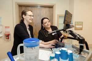 Two healthcare professionals stand at a medical cart, looking at a computer monitor in a clinical setting.