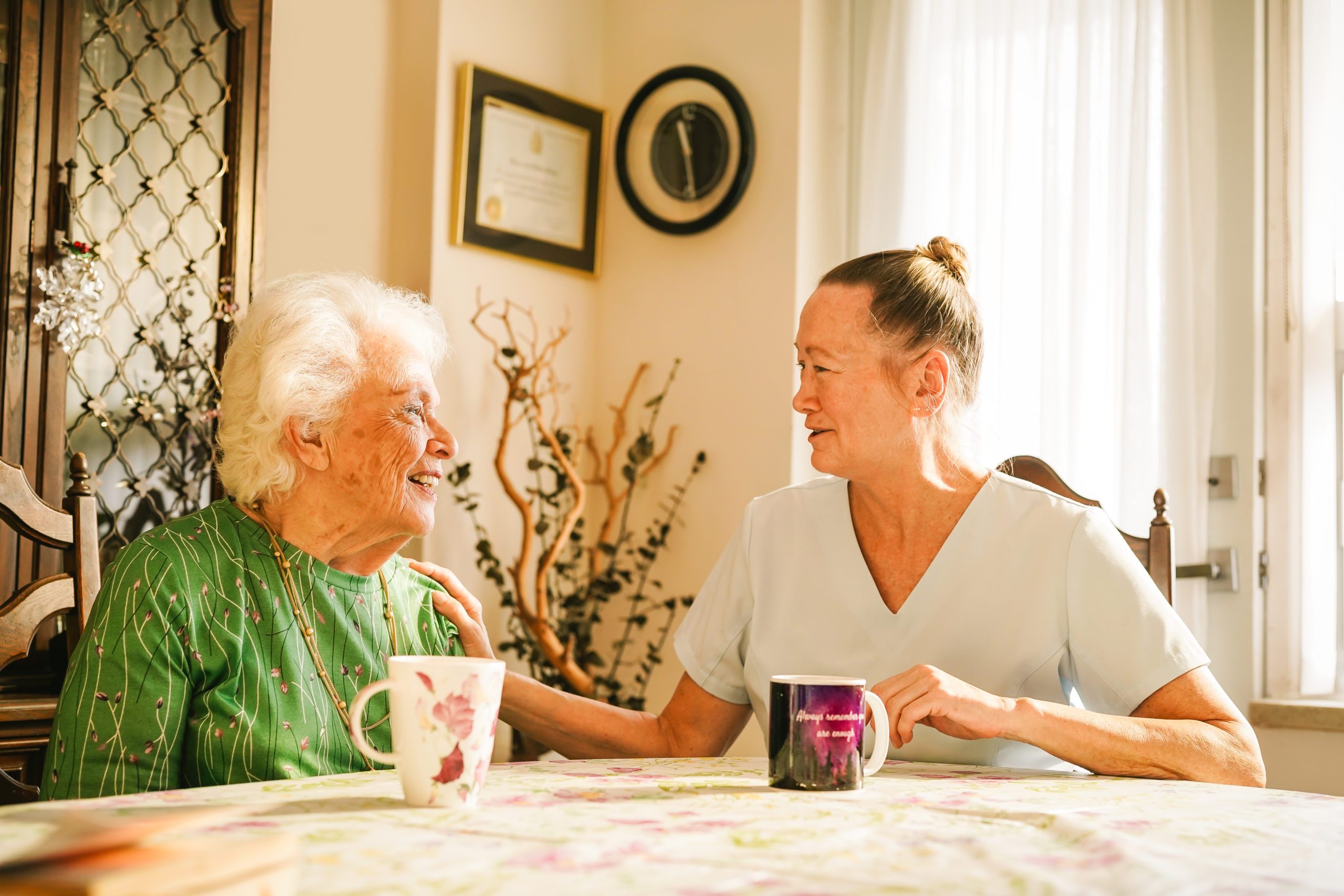 An elderly woman and a caregiver sit at a table with mugs, smiling and talking in a sunlit room.