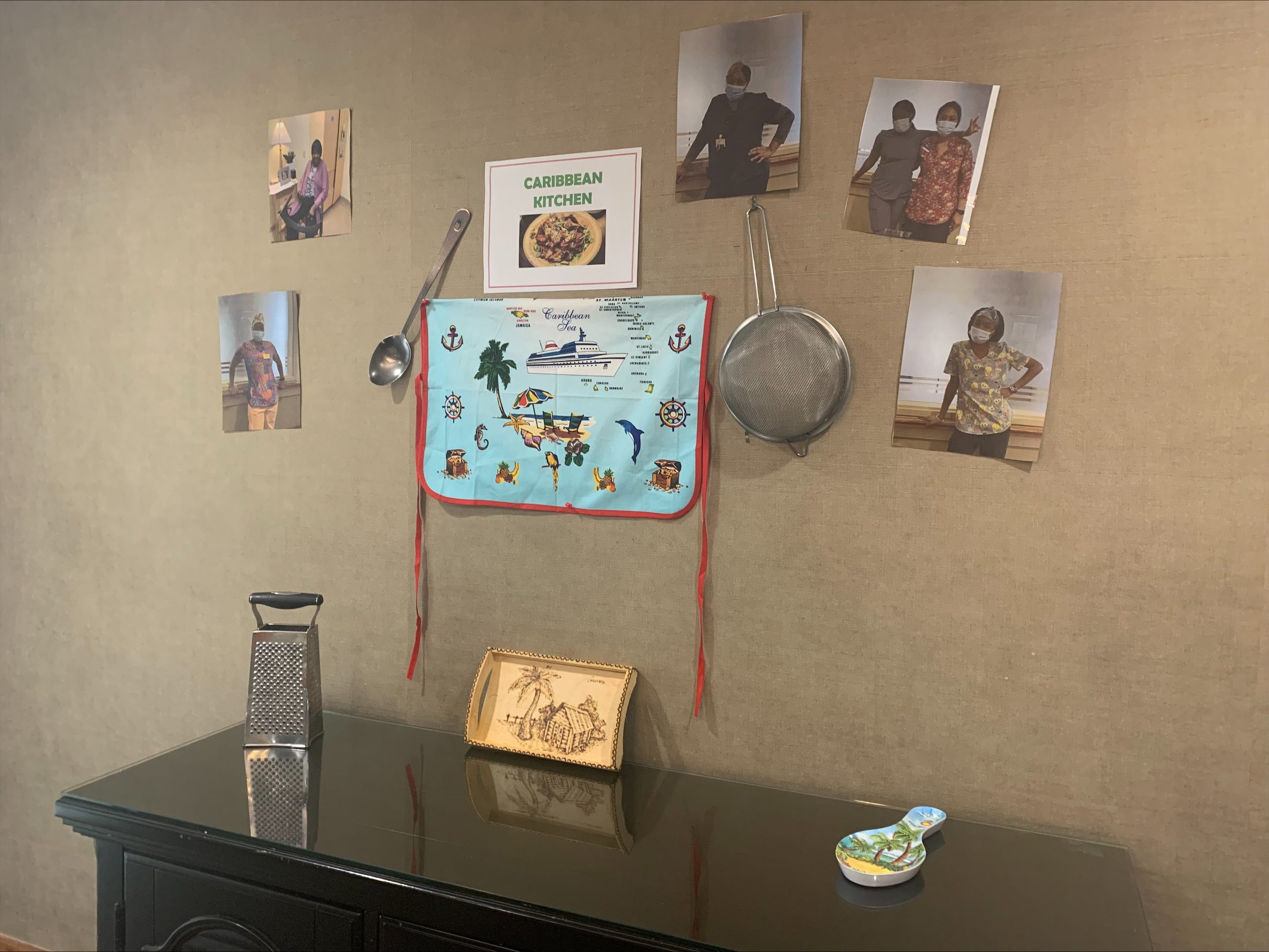 A wall display features a “Caribbean Kitchen” sign, photos of people, kitchen utensils, a decorative map, and a grater on a black table.