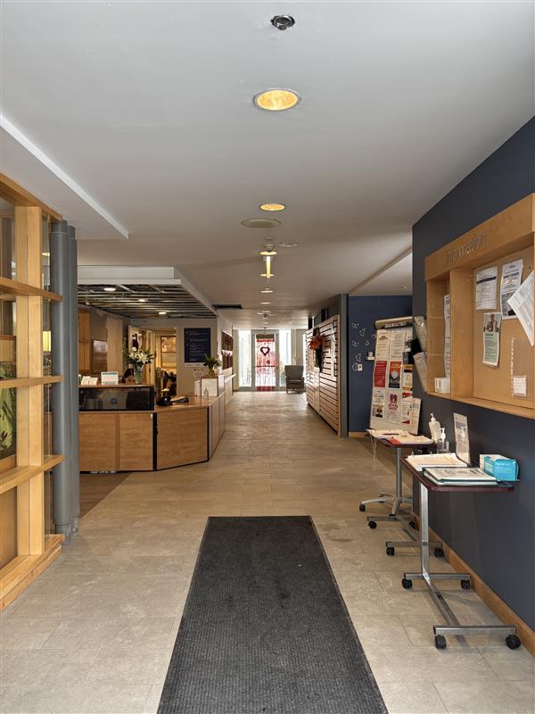 A hallway with tiled floors leads to a reception desk on the left and a bulletin board with flyers on the right; a mat runs along the center.