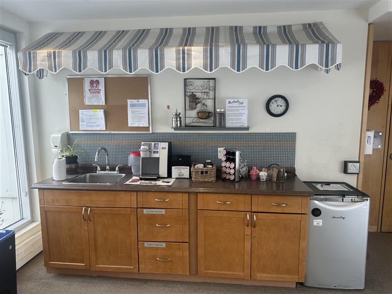 Coffee station with wooden cabinets, a small fridge, sink, coffee machines, condiments, bulletin board, and wall clock under a striped awning.