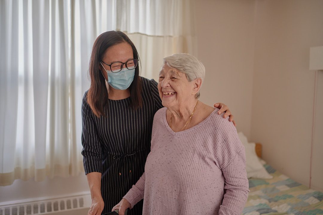 A younger woman wearing a face mask stands next to and supports an older woman in a bedroom, both smiling.