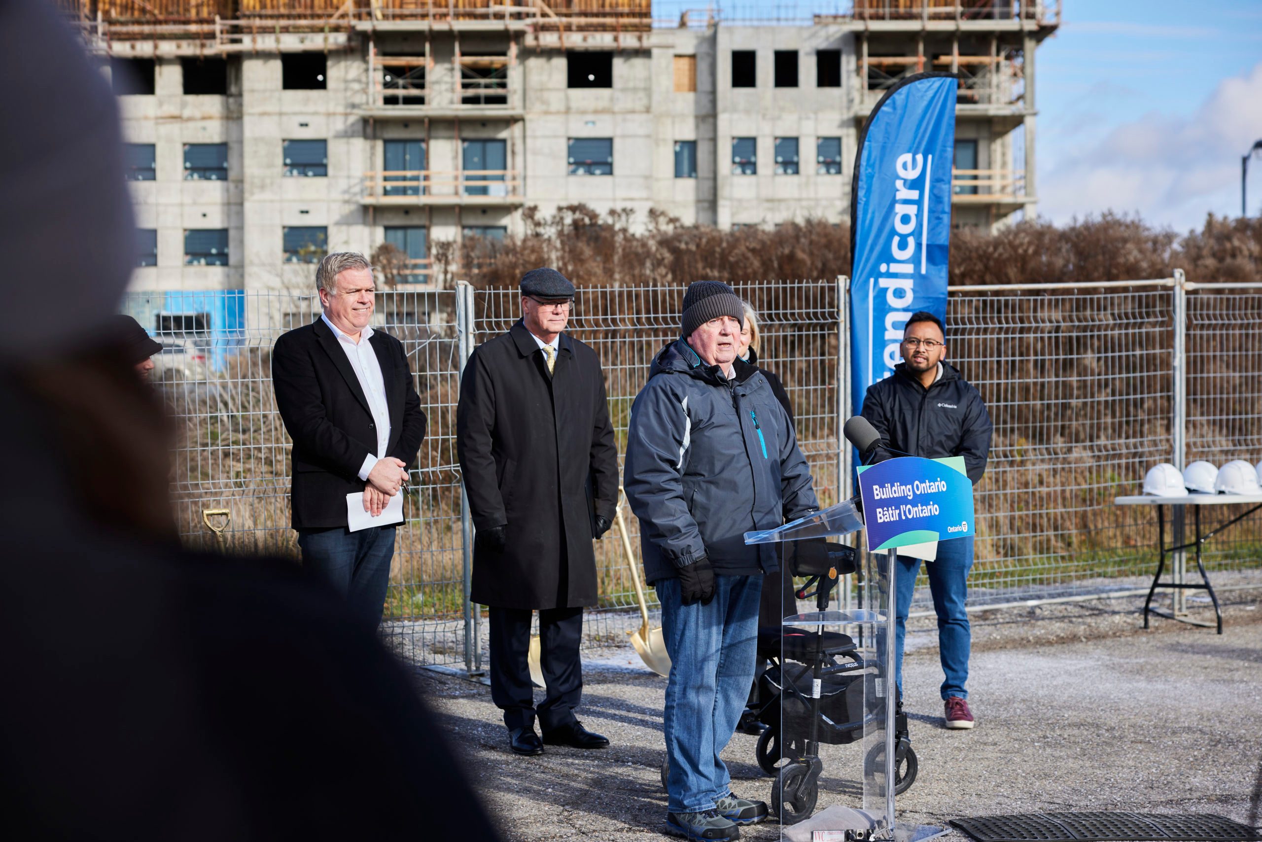 A speaker addresses an outdoor press event at a construction site, with three people standing behind and construction in the background.