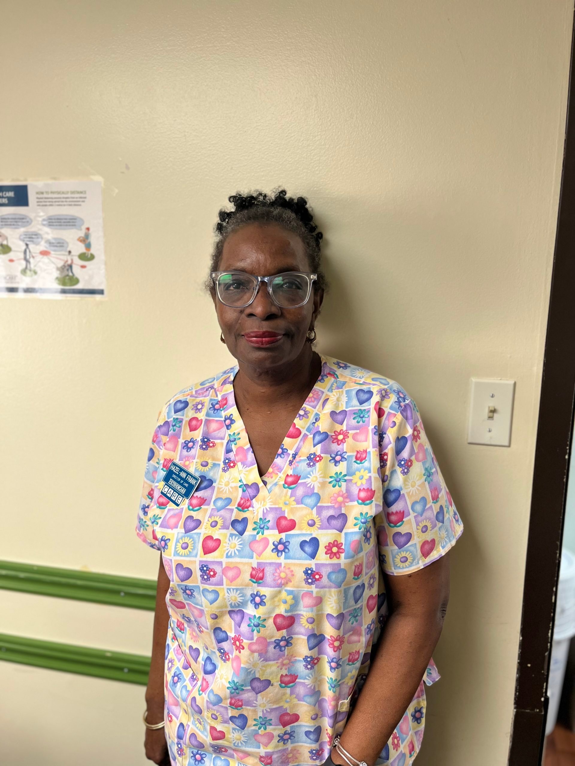A woman in colorful scrubs with a floral and heart pattern stands against a beige wall in an indoor setting.