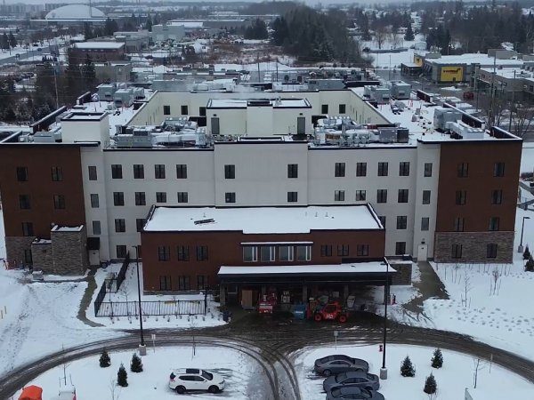 Aerial view of a multi-story building with a snow-covered roof and surrounding ground, machinery near the entrance, and cars parked in front.