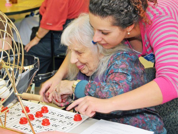 A young woman assists an elderly woman in placing markers on a bingo card at a table, with a bingo cage visible nearby.