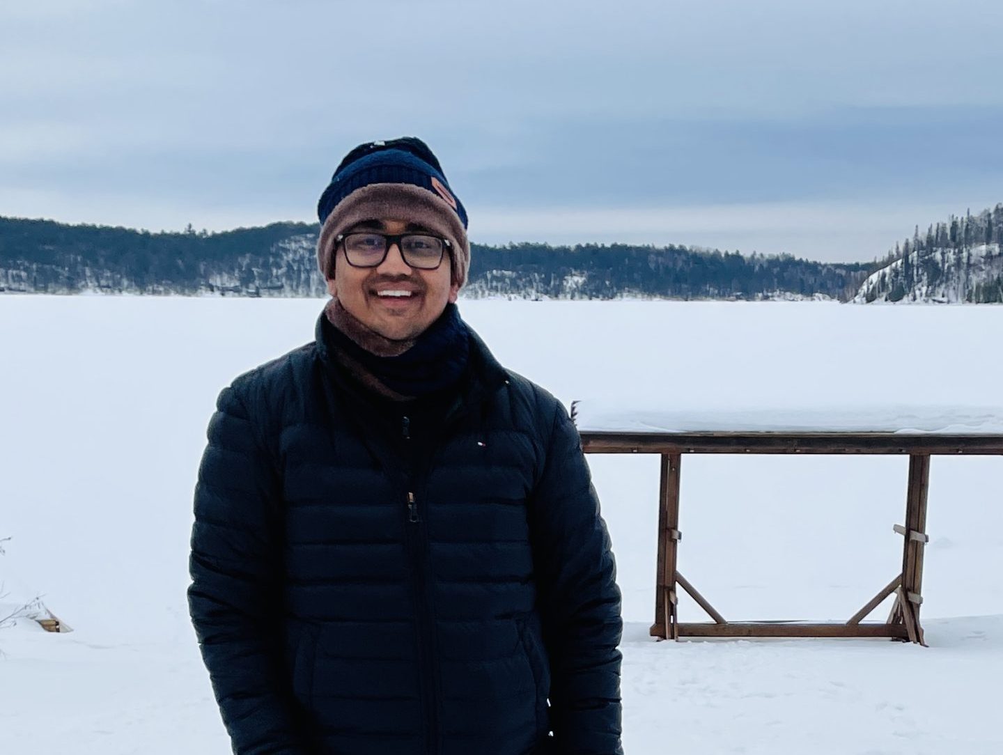 A man stands on snow wearing winter clothes and glasses, smiling, with a golden retriever lying at his feet; a frozen lake and snow-covered hills are in the background.
