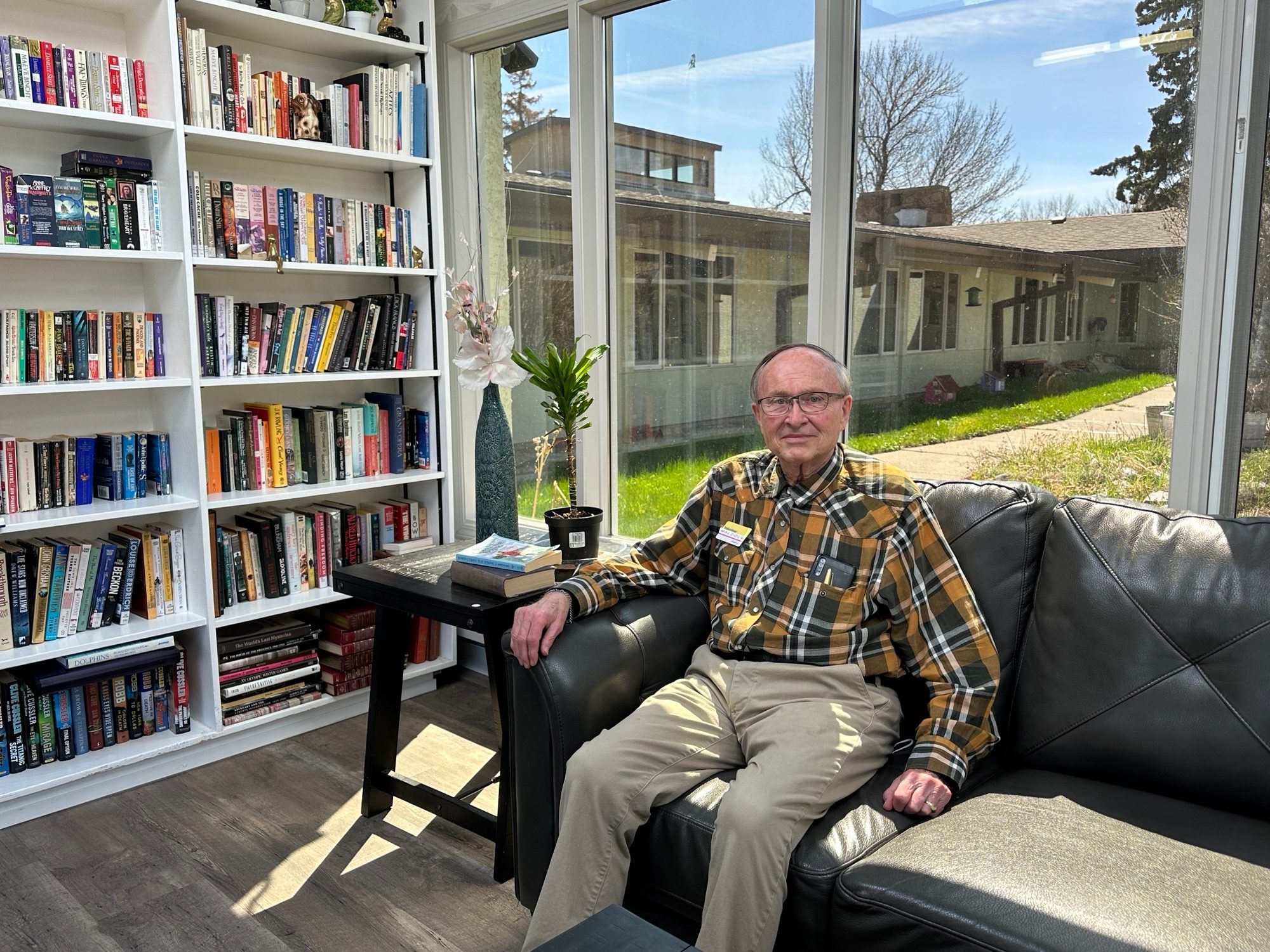 An older man sits on a black leather sofa in a sunlit room with large windows, next to a bookshelf filled with books and a small table with a plant.