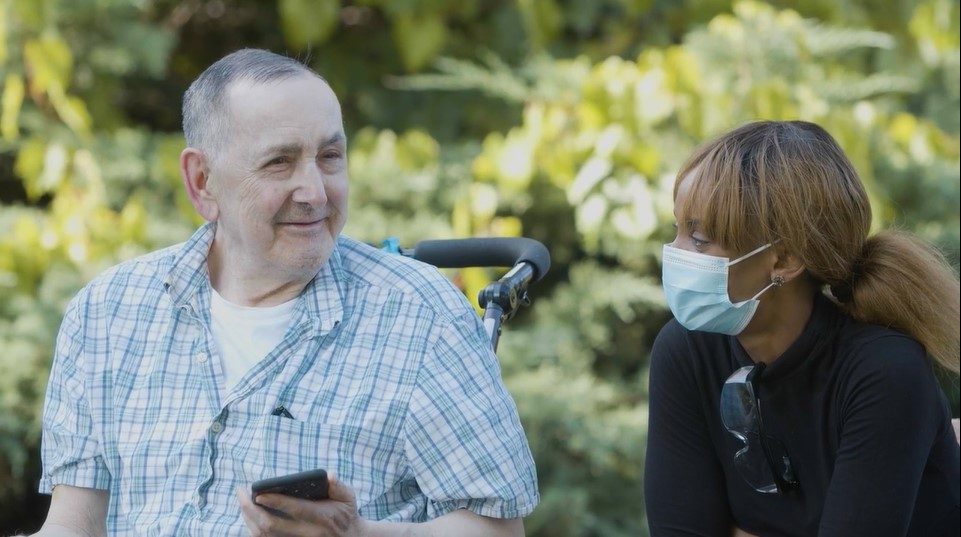 An older man holding a smartphone sits outside next to a woman wearing a face mask; both appear to be talking. Lush greenery is in the background.