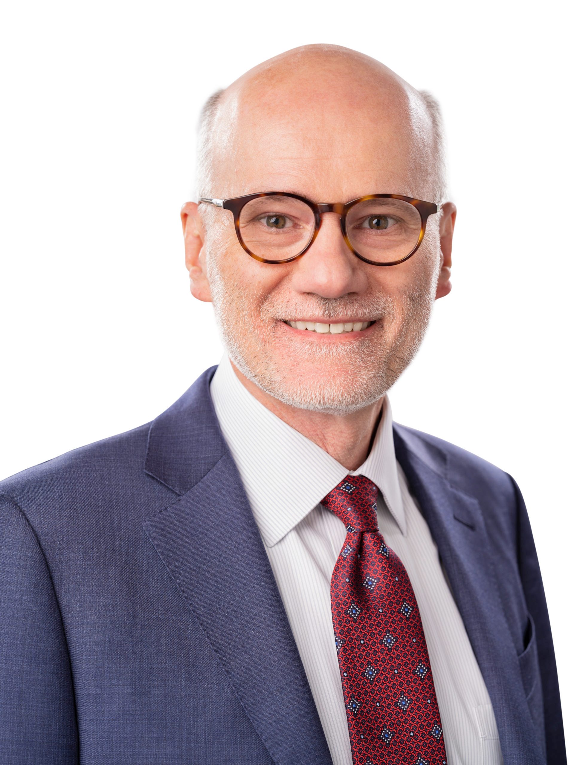 A middle-aged man with glasses, short gray hair, and a beard, wearing a blue suit, white shirt, and red patterned tie, poses against a white background.