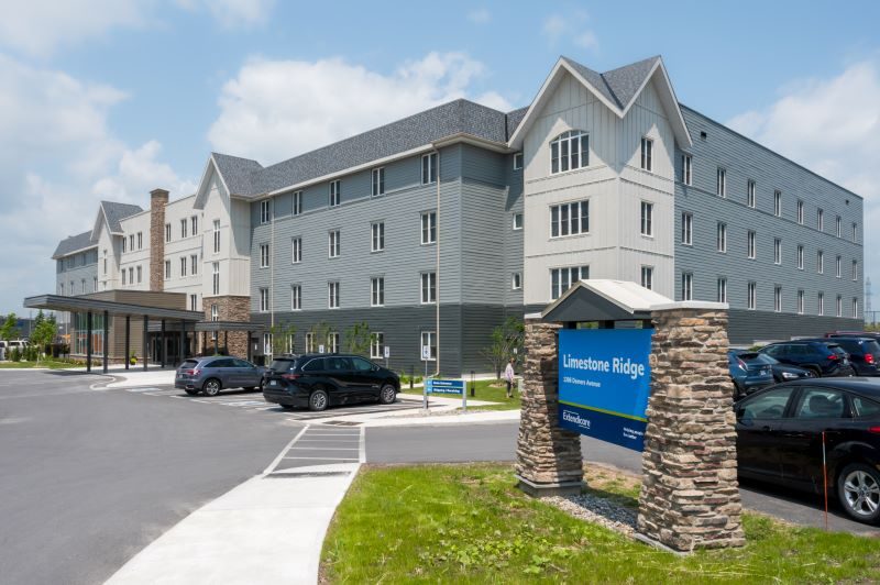 A modern three-story building with gray siding and stone accents, a parking lot with cars, and a blue sign reading "Limestone Ridge" in front.