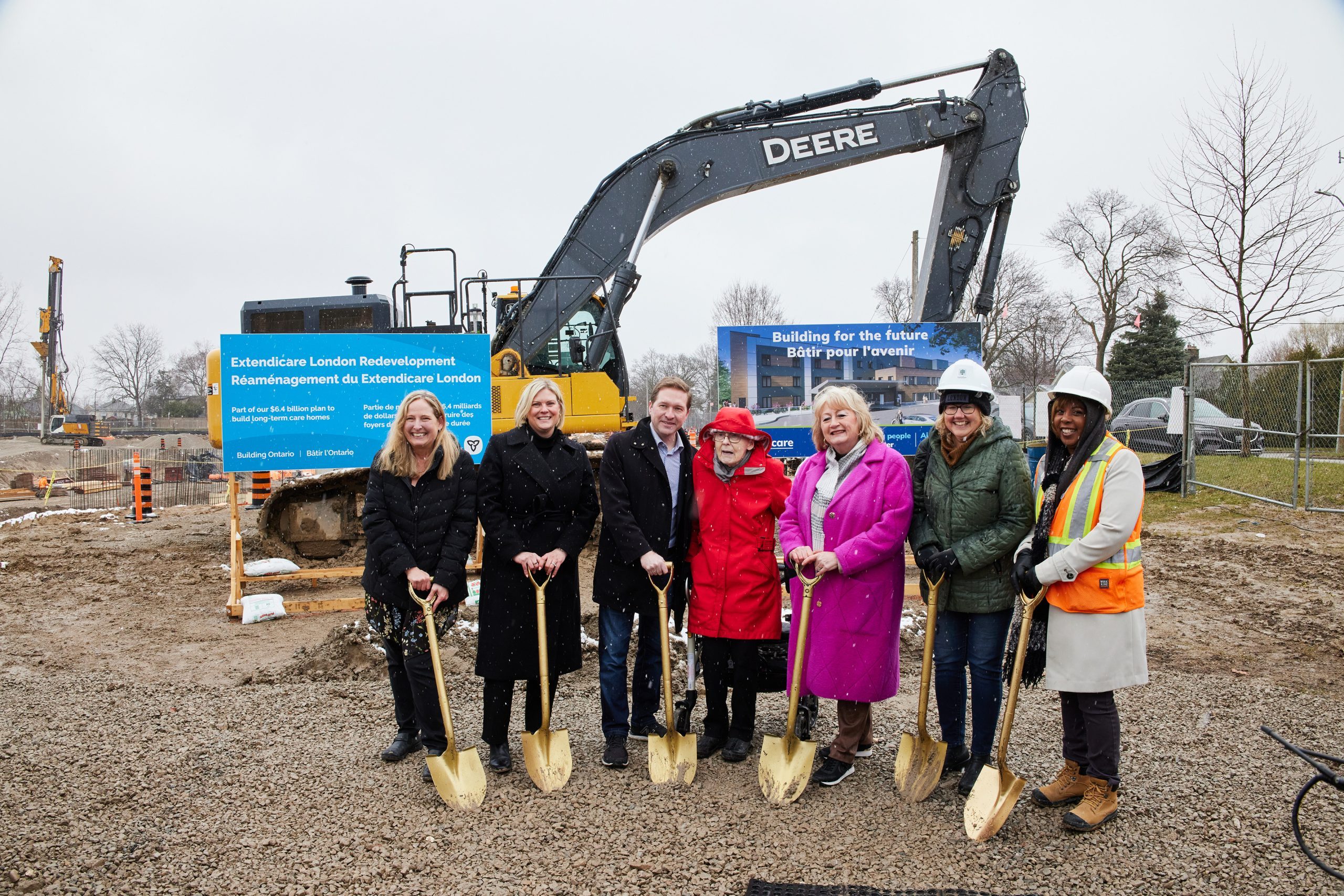 A group of seven people in coats and helmets pose with shovels at a construction site, standing in front of a large excavator and informational signs.