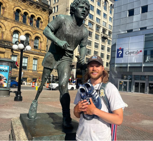A man stands smiling in front of a running statue, holding a pair of running shoes in an urban plaza with historic and modern buildings in the background.