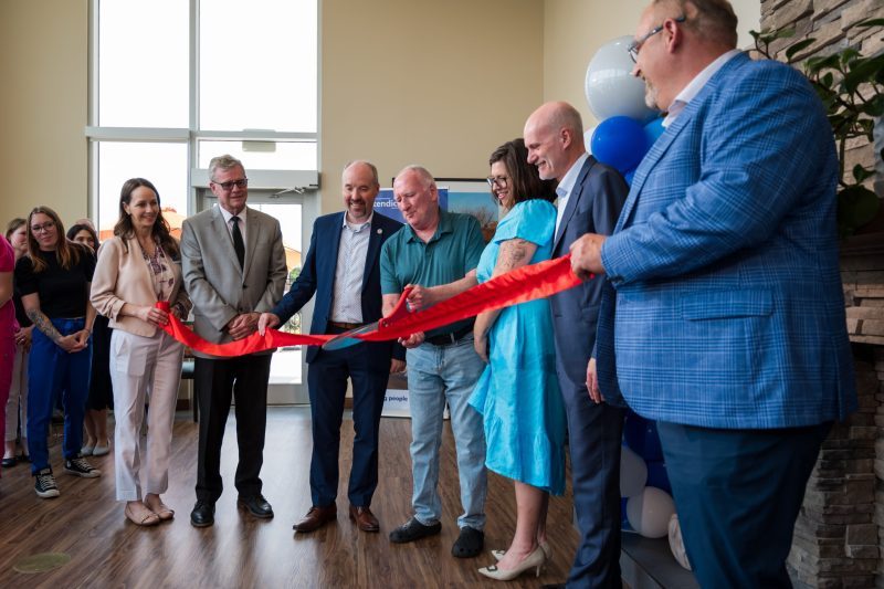 A group of people stand indoors as one person cuts a red ribbon with large scissors during a ribbon-cutting ceremony. Some attendees are smiling and watching.