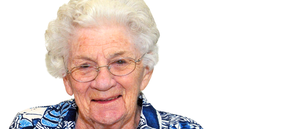 An elderly woman with white hair and glasses smiles at the camera, wearing a patterned blue and white shirt against a plain light background.