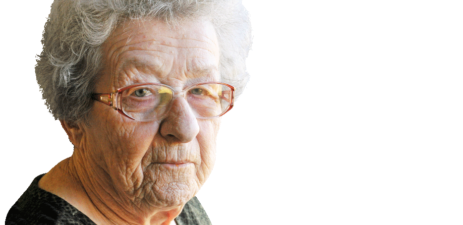 Elderly woman with short gray hair and glasses looks toward the camera, wearing a dark top and neutral expression against a light background.