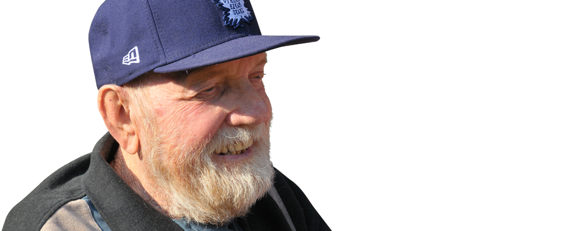 Elderly man with a white beard wearing a blue Toronto Maple Leafs cap and a dark jacket, smiling and looking to the side against a plain background.