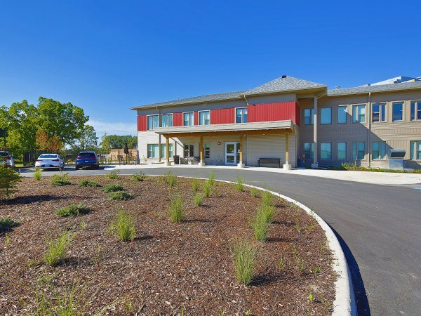 A modern two-story building with red and beige accents, a covered entrance, and a circular driveway surrounded by landscaped shrubs and parked cars.