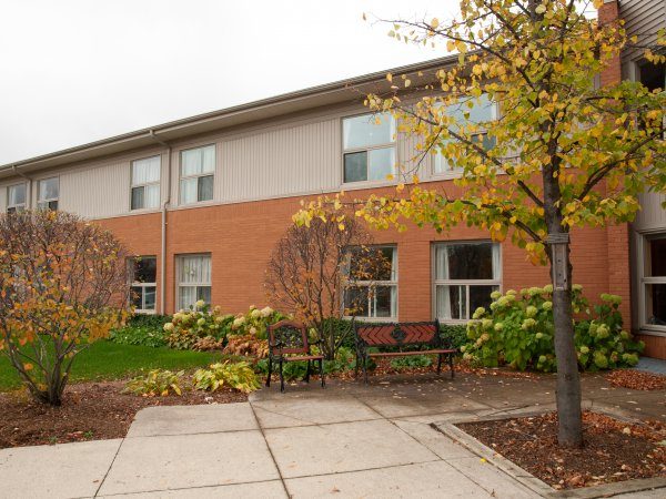 A two-story brick building with multiple windows, a bench, trees with autumn leaves, and a concrete walkway in the foreground.