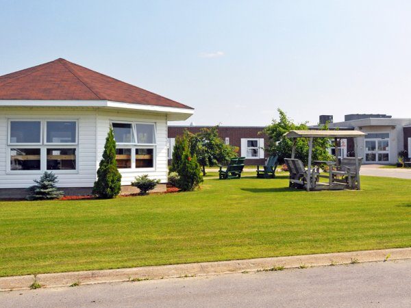 A well-kept lawn with a small white building, benches, and a gazebo in front of a larger complex under a clear sky.