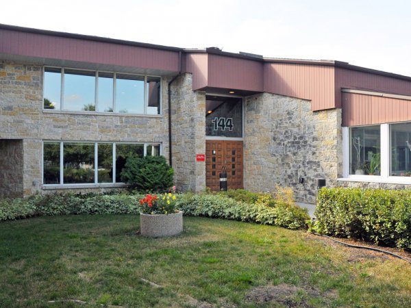 A stone and wood office building with large windows, a wooden door numbered 144, and a landscaped lawn with a concrete planter of flowers in front.