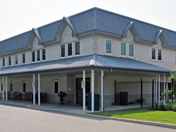 A two-story building with a gray metal roof and white exterior walls, featuring multiple windows and a covered porch supported by white columns.