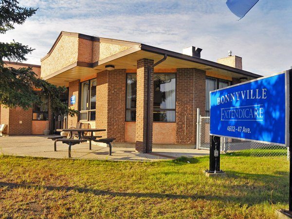 A brick building with large windows and a blue sign reading "Bonnyville Extendicare, 4602-47 Ave." sits under a partly cloudy sky. A picnic table is visible outside.