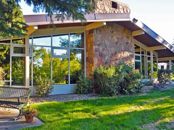 A modern building with large glass windows, a stone wall, and a sloping roof sits surrounded by bushes, trees, and a grassy lawn.
